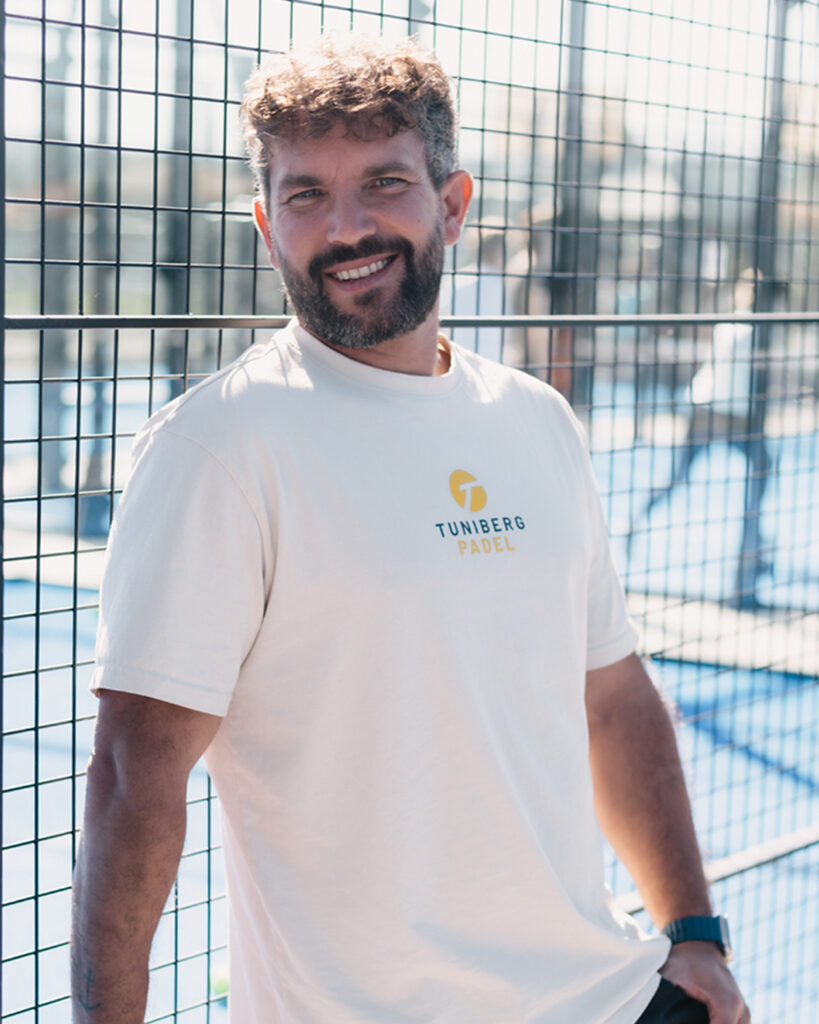 Ein Mann lehnt lächelnd an einem Zaun auf einem Padel-Tennisplatz, trägt ein helles T-Shirt mit „Tunberg Padel“-Logo.