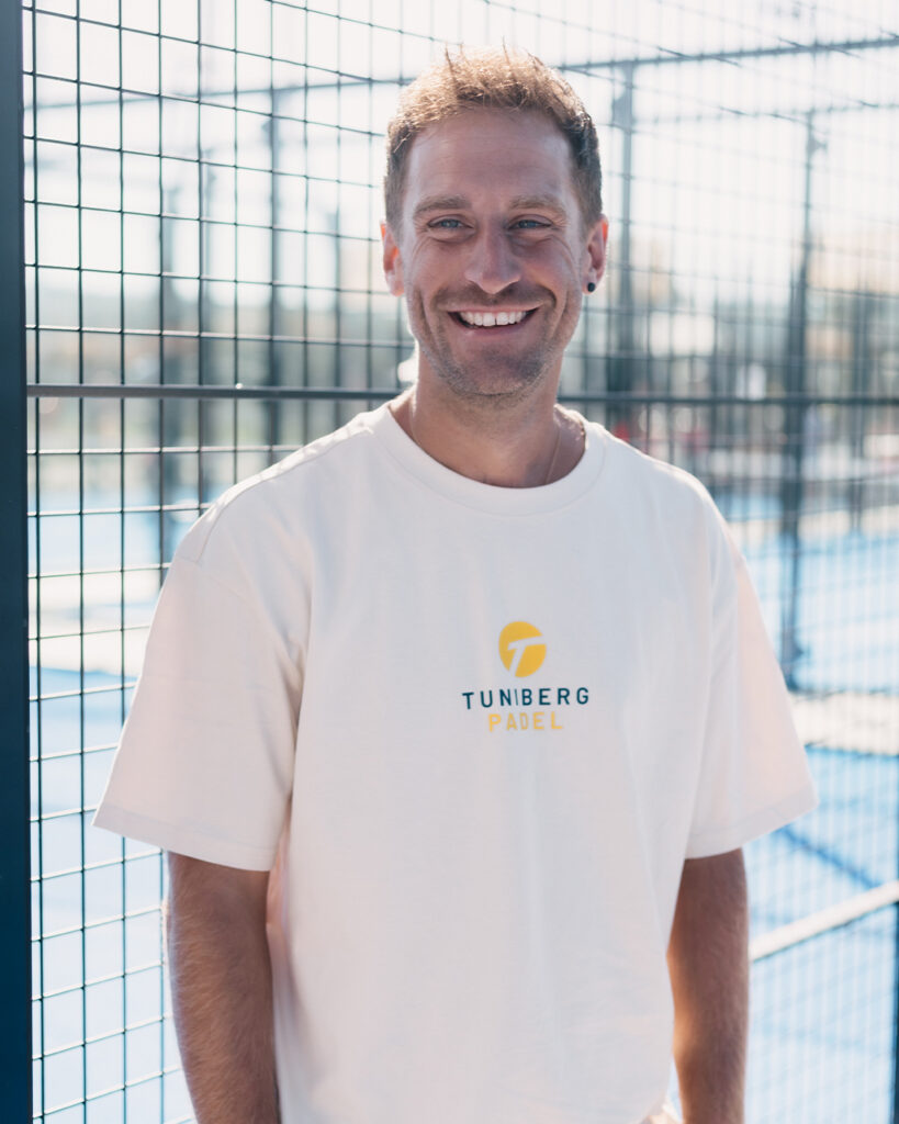 Ein Mann steht lächelnd vor einem Gitterzaun auf einem Padelplatz und trägt ein weißes T-Shirt mit der Aufschrift „Tunberg Padel“.
