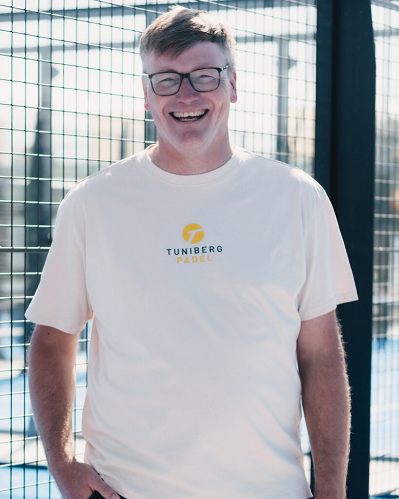 Ein lächelnder Mann mit Brille steht draußen an einem Padelplatz und trägt ein weißes "Tuniberg Padel"-T-Shirt.