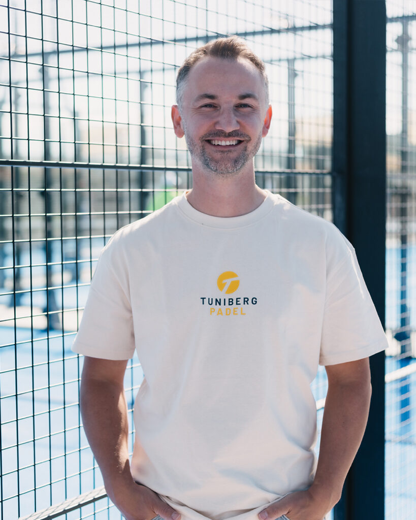 Ein lächelnder Mann steht vor einem Tennisplatzzaun und trägt ein weißes T-Shirt mit der Aufschrift „Tuniberg Padel“.